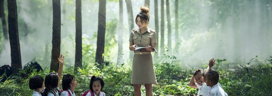 This is an image of a school where the teacher is teaching several little children outdoors amidst trees